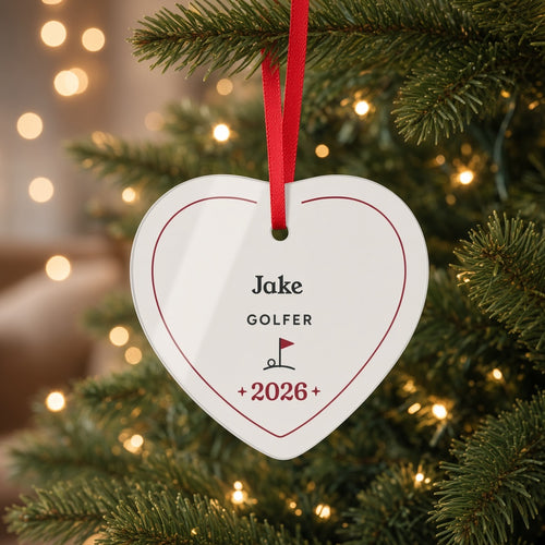 Personalized heart acrylic Christmas ornament with the name Jake, GOLFER, a golf flag and 2026, hanging by a red ribbon on a lit Christmas tree